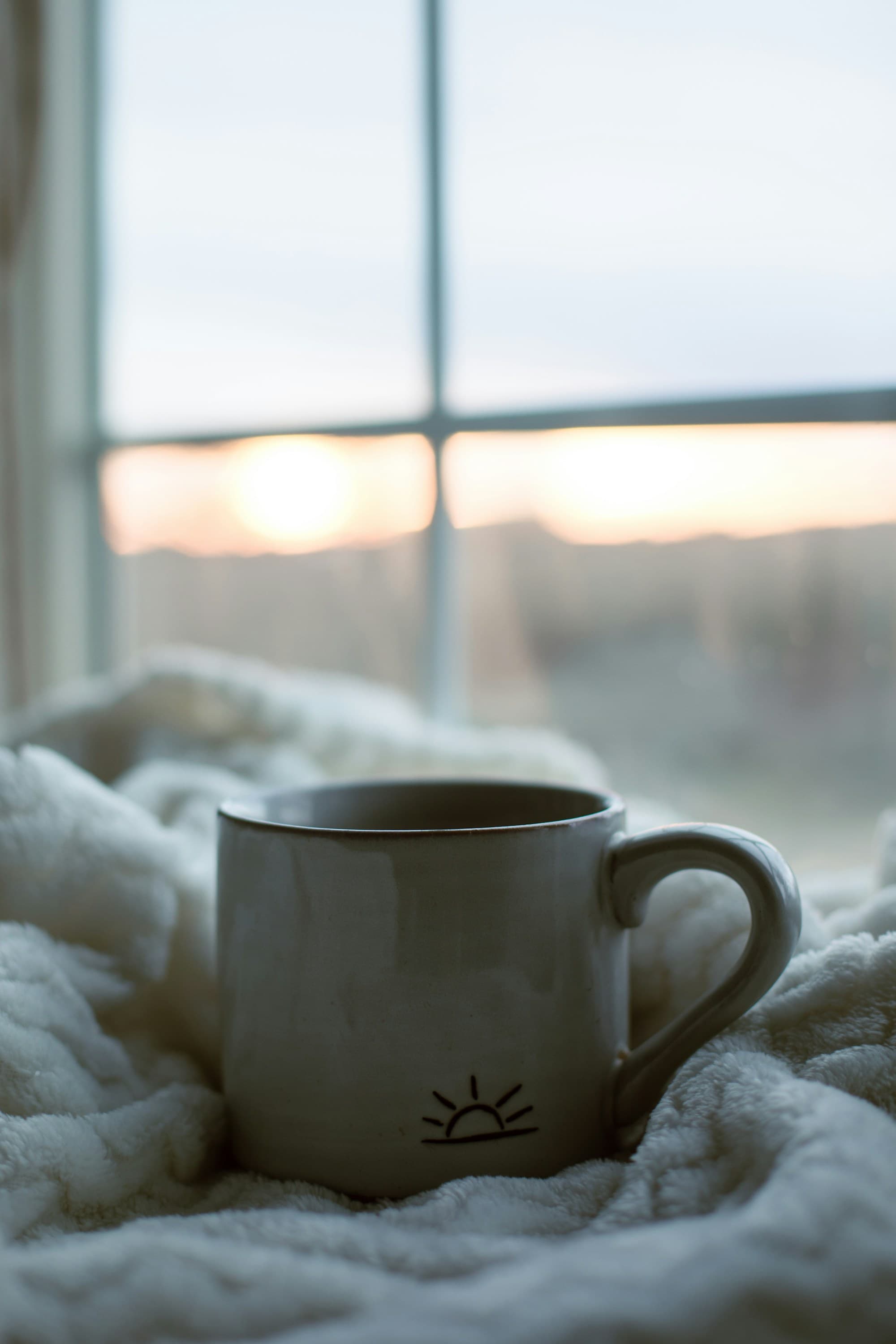 A handmade ceramic mug with a rising-sun icon, nestled in a soft blanket, with the dawn rising through the window beyond — Daybreak Therapy