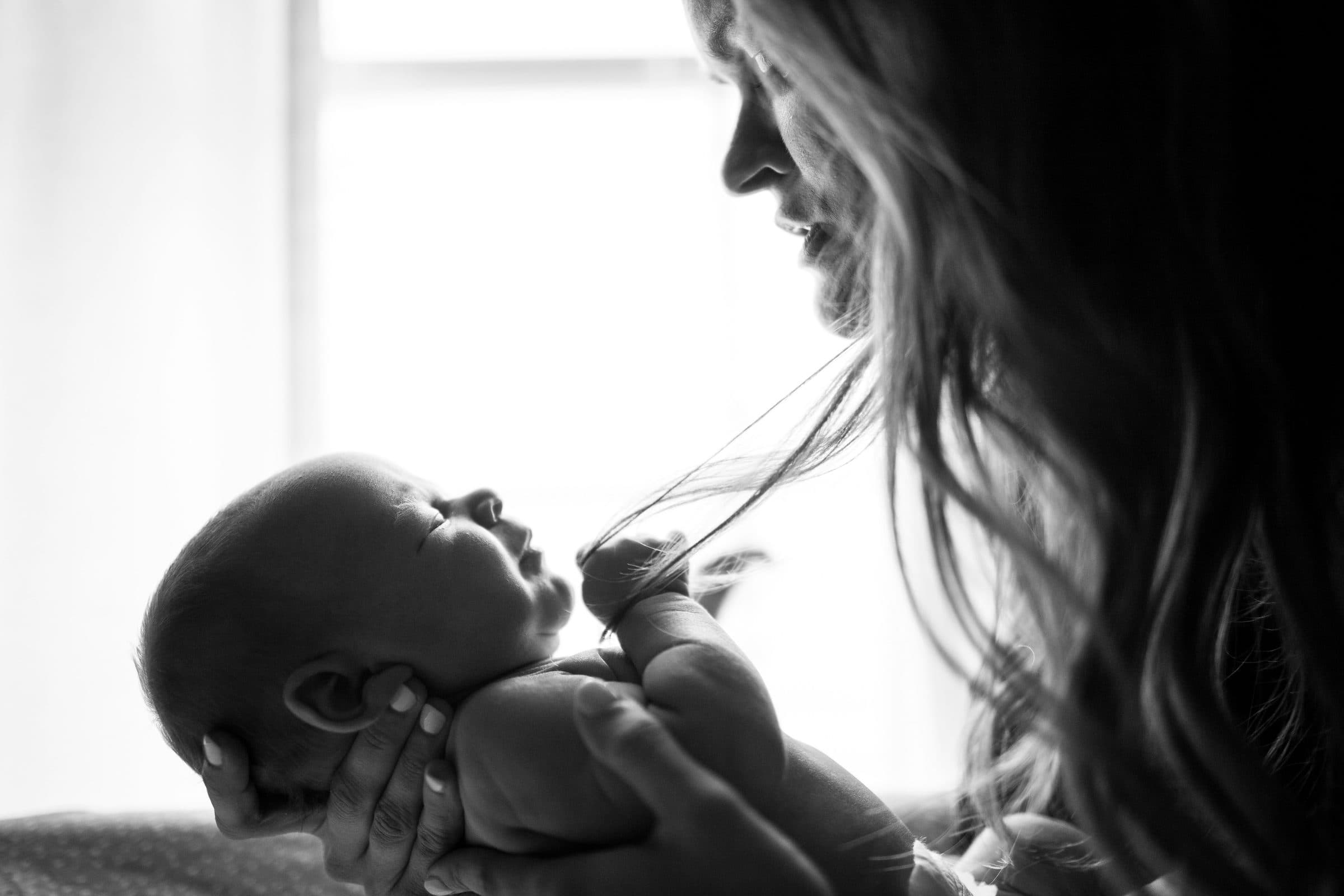 A mother in profile looking down at her newborn, backlit by a window, in black and white