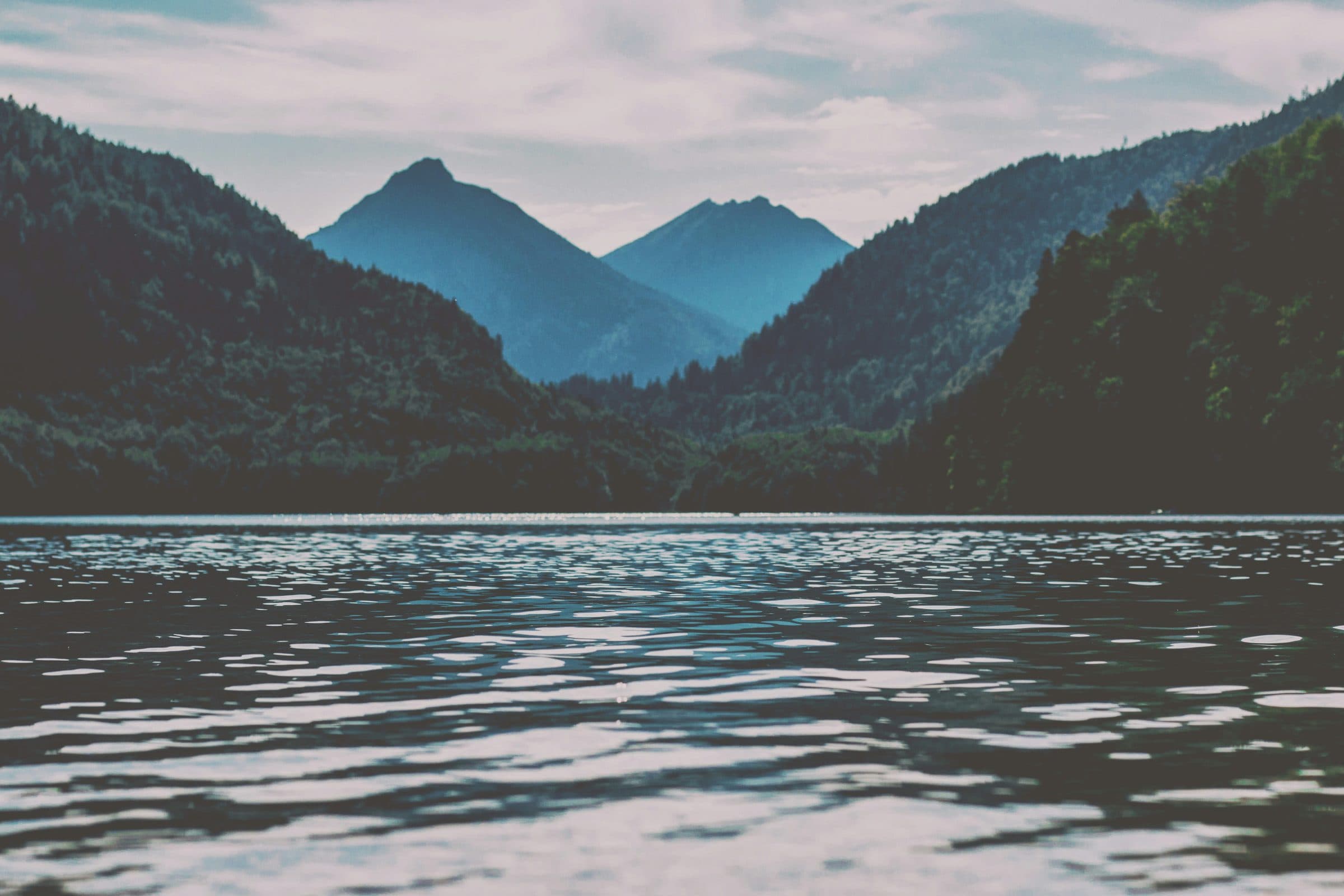 Still lake water in the foreground with forested hillsides and blue mountain peaks beyond